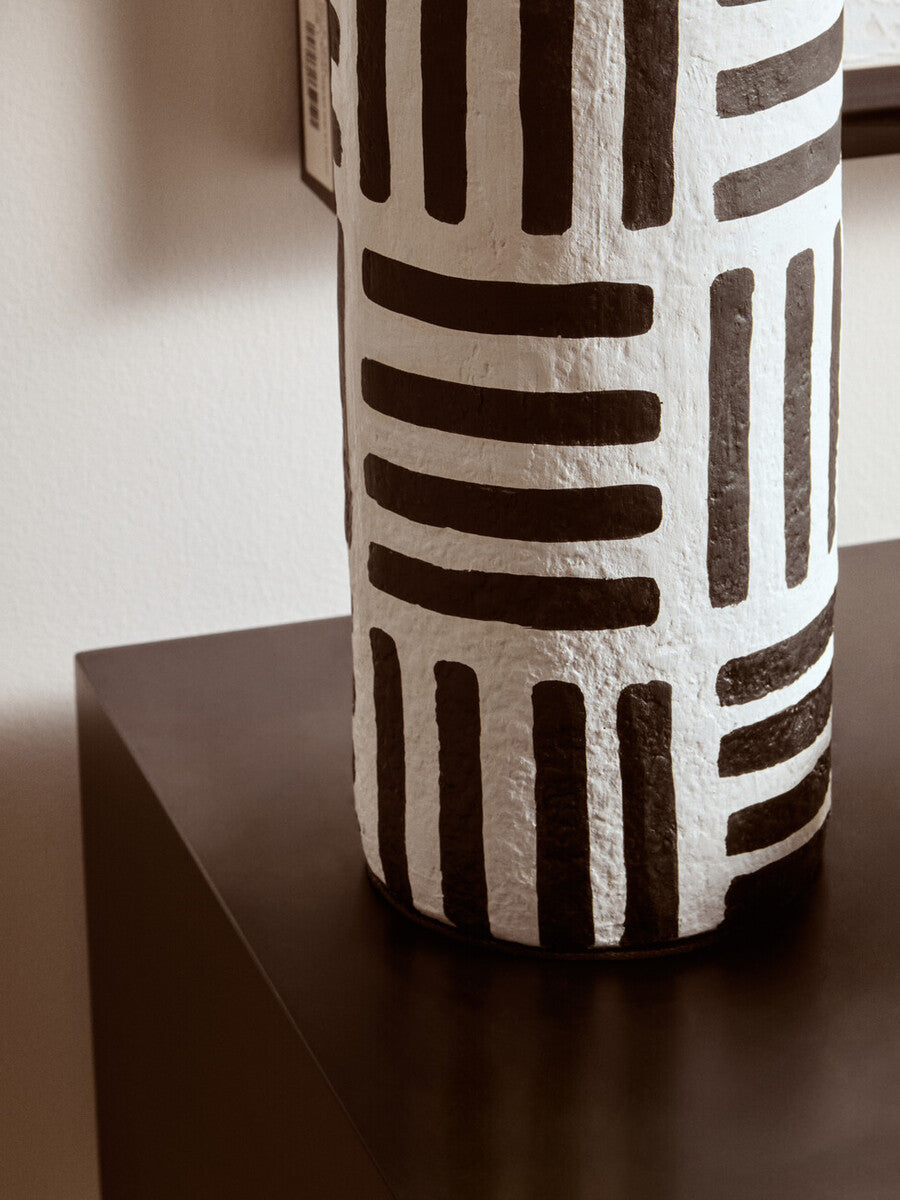 A white papier mache table lamp with bold black stripes and a textured cylindrical ceramic base with geometric lines
