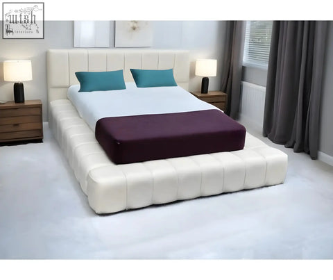 Cream-colored tufted cloud bed with padded headboard and foot end, featuring teal pillows and purple throw blanket on bed frame