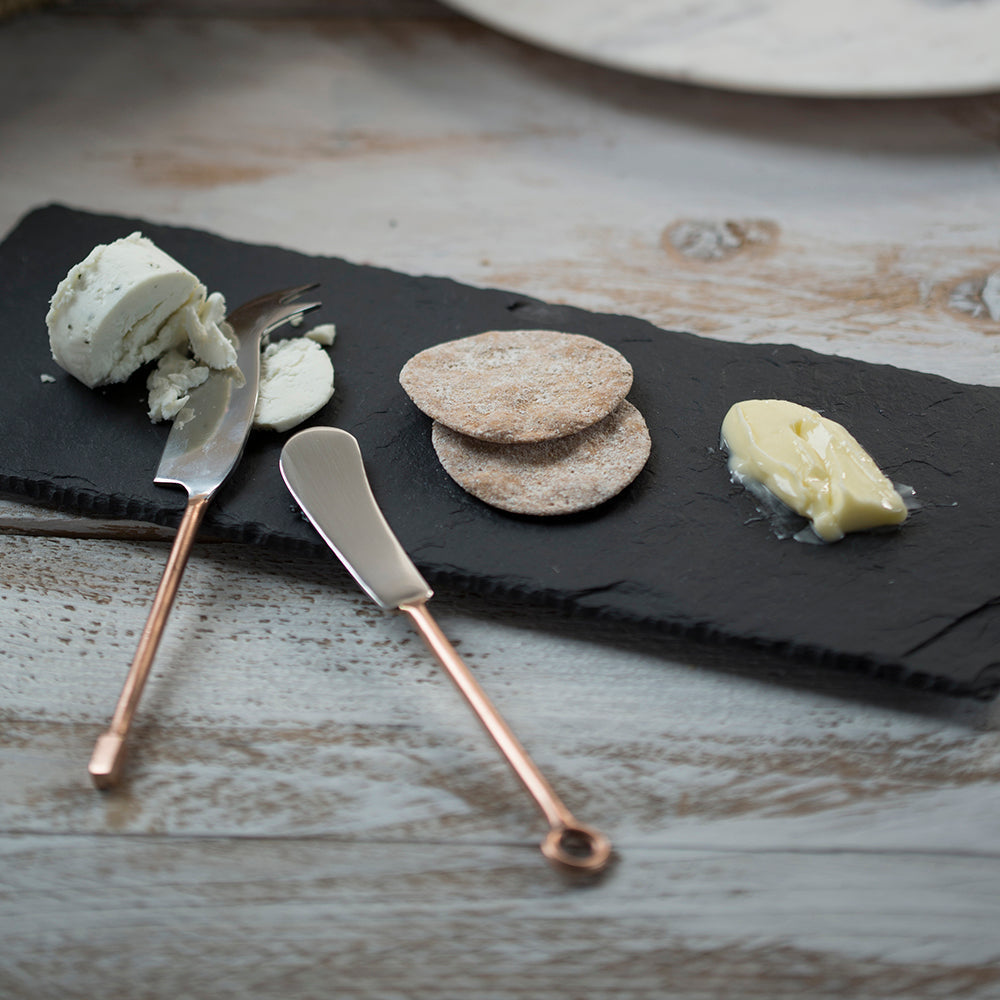 A set of 2 copper cheese knives with copper handles and polished metal blades resting on a dark slate board