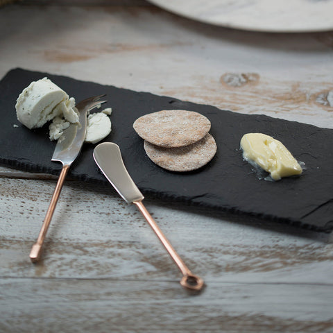 A set of 2 copper cheese knives with copper handles and polished metal blades resting on a dark slate board