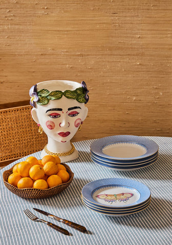 A white ceramic vase shaped like a woman face with painted features and green crown beside cornflower blue lace butterfly plates