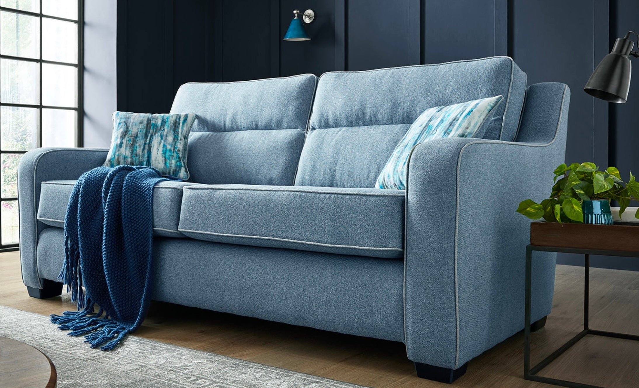 Light blue textured fabric cornwall sofa with shaped square arms, two patterned pillows, and navy knit blanket draped