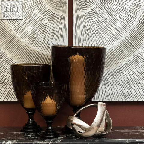 Dark amber cortina large honeycomb glass candle holder with hammered texture and black base next to silver sculpture