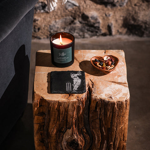 A lit amber glass candle with a dark green sibra label featuring cricket bats on a sleek black single slate coaster