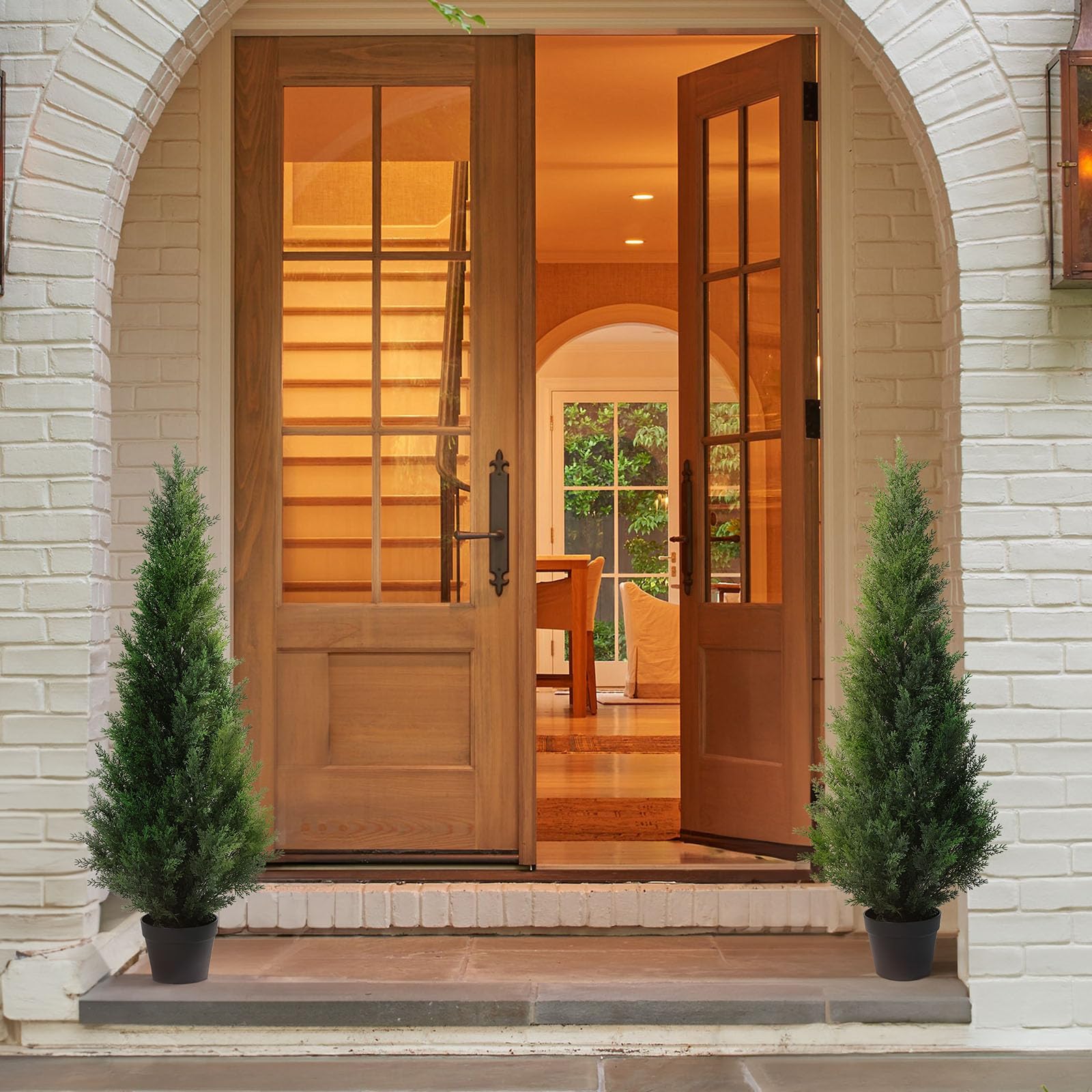 Warm-toned wooden double doors with glass panes and dark handles framed by white brick and artificial cedar tree decor