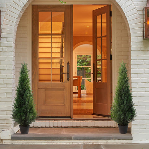 Warm-toned wooden double doors with glass panes and dark handles framed by white brick and artificial cedar tree decor