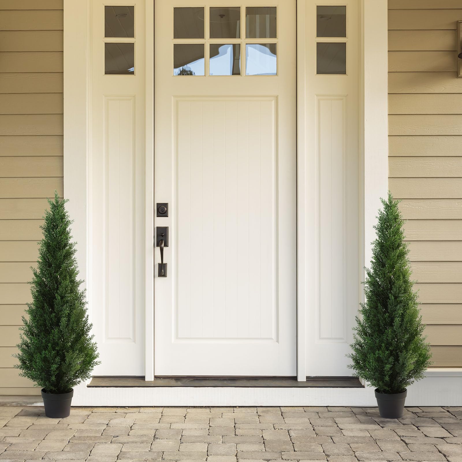 White front door with black handle flanked by two tall green artificial cedar trees in black pots for porch decor