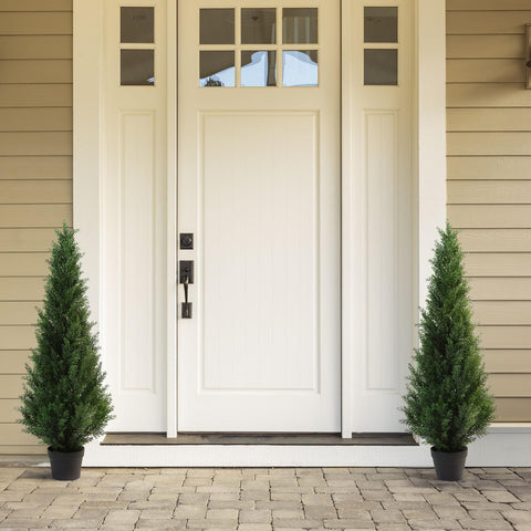 White front door with black handle flanked by two tall green artificial cedar trees in black pots for porch decor