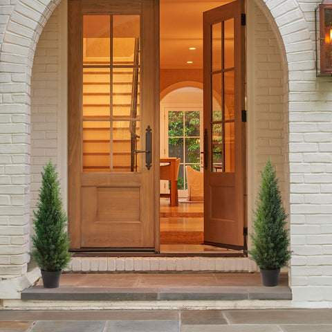 Warm-toned wooden double doors with glass panes and dark metal hardware framed by white brick archway near artificial cedar tree pots