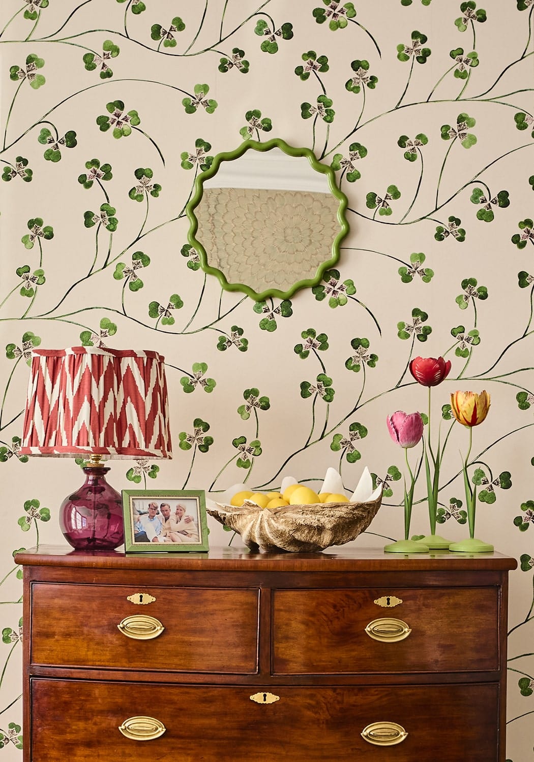 A green scalloped mirror with a decorative frame hangs above a wooden dresser near a dahlia pink tulip candleholder tole