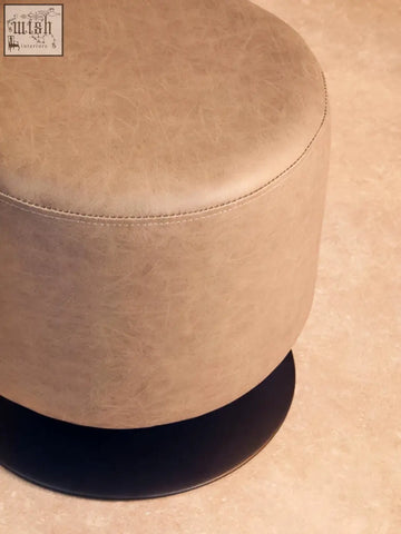 Beige round ottoman with ash grey faux leather upholstery and a black circular base as a stylish faux leather pouffe