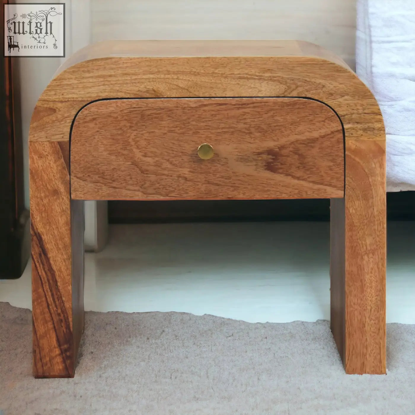 A sleek curved solid mango wood darcy bedside table with a single drawer and brass round knob