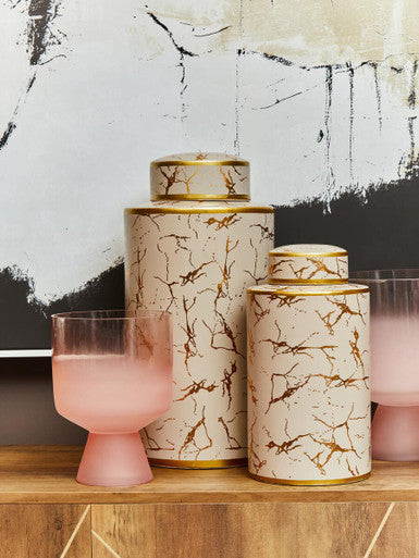 Two daria large ceramic jars with cream marble patterns and gold accents next to a matching pink frosted glass