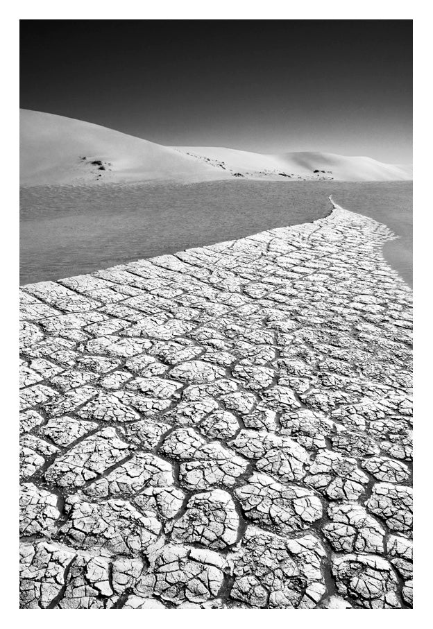 Fine art photo by david ballam showing cracked desiccated earth with a winding path to dunes under stark sky