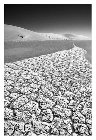 Fine art photo by david ballam showing cracked desiccated earth with a winding path to dunes under stark sky