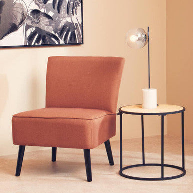 A cozy rust-colored armchair with sleek black legs next to a depok rattan and metal round side table