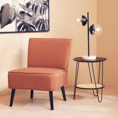 A cozy orange armchair with sleek black legs next to a depok rattan side table with hairpin legs and lower shelf