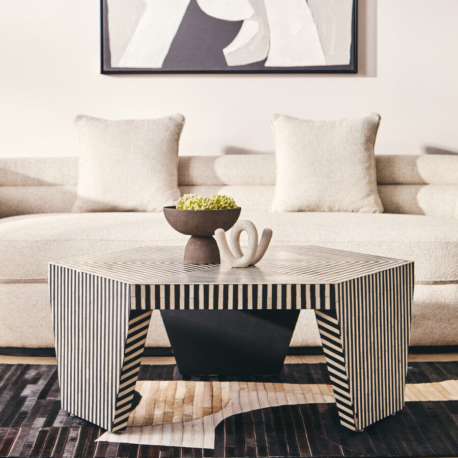 Hexagonal coffee table with black and white stripes and sleek geometric legs next to a textured grey pedestal bowl