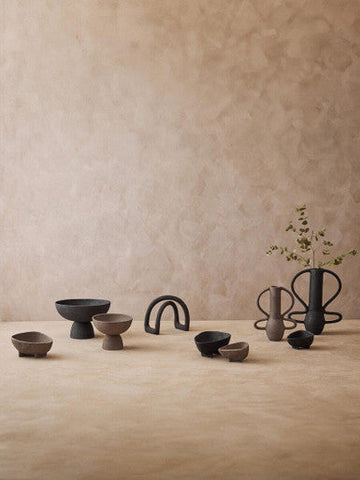 A sleek matte black ceramic vase with curved handles and slender neck beside a textured grey pedestal bowl