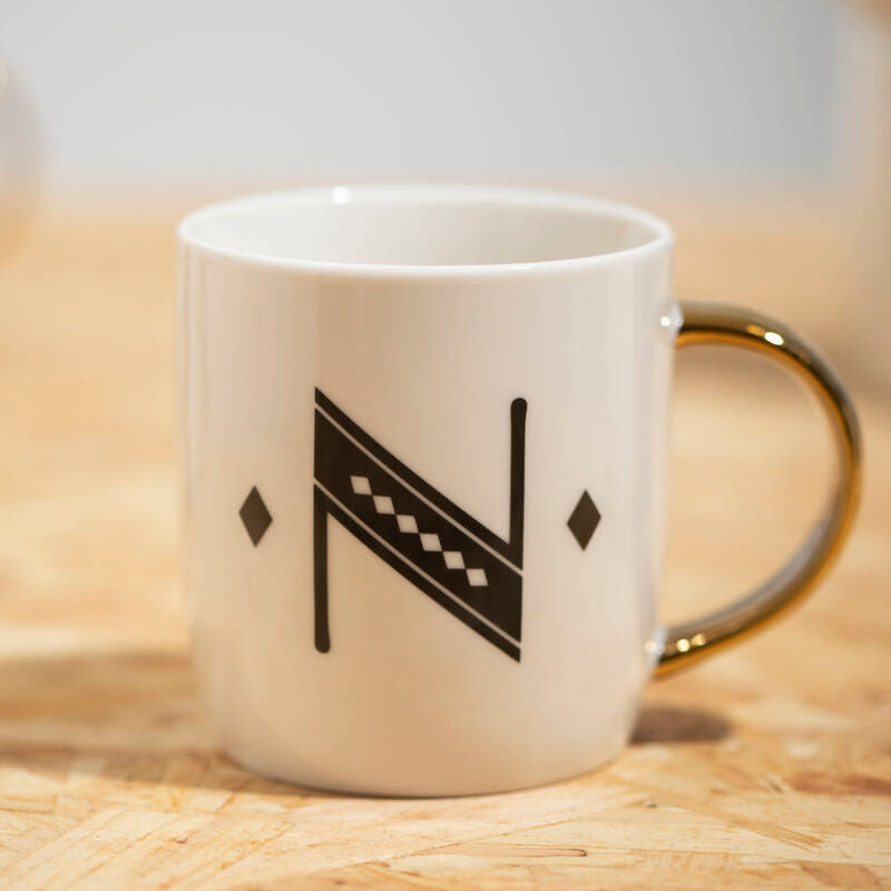 White ceramic mug with gold handle and black geometric n design featuring diamond deco accents in deco white style