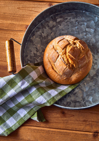 Golden brown loaf on a green checkered cloth inside a three round galvanised steel tray with wooden handles