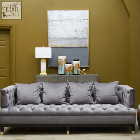 Gray tufted velvet sofa with dark wood legs and gold feet next to edith table lamp with white marble base and gold accents