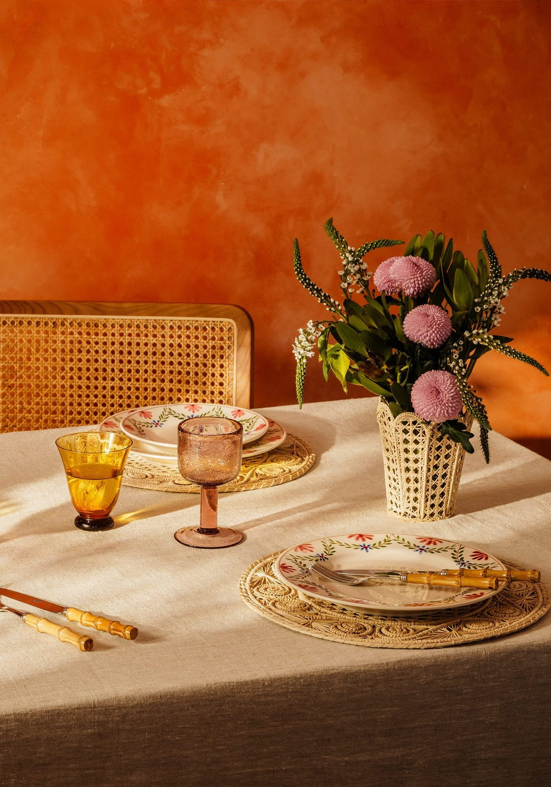 A cream-colored woven vase with a geometric pattern holding pink and white flowers shown beside eleanor wine glass violet