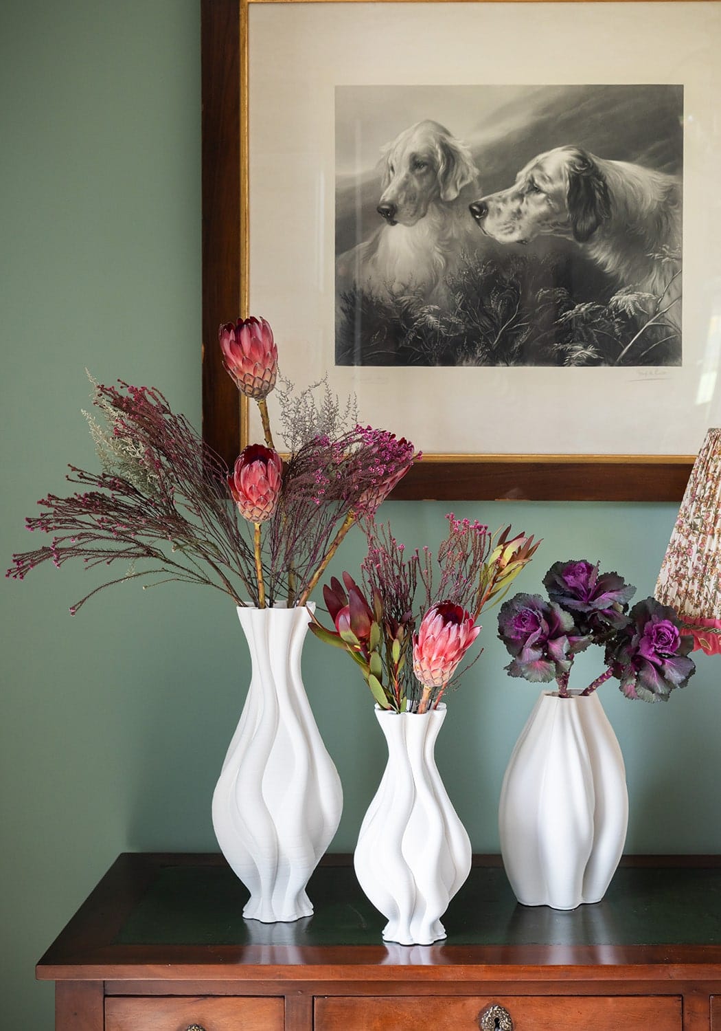 Three white ceramic vases with wavy designs hold pink proteas and purple cabbage in emilie white ceramic vase