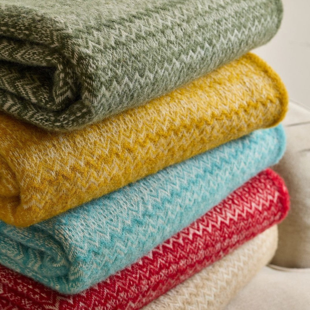 Stack of fair isle pure new wool throw blankets in olive green mustard yellow teal blue and red with textured zigzag pattern