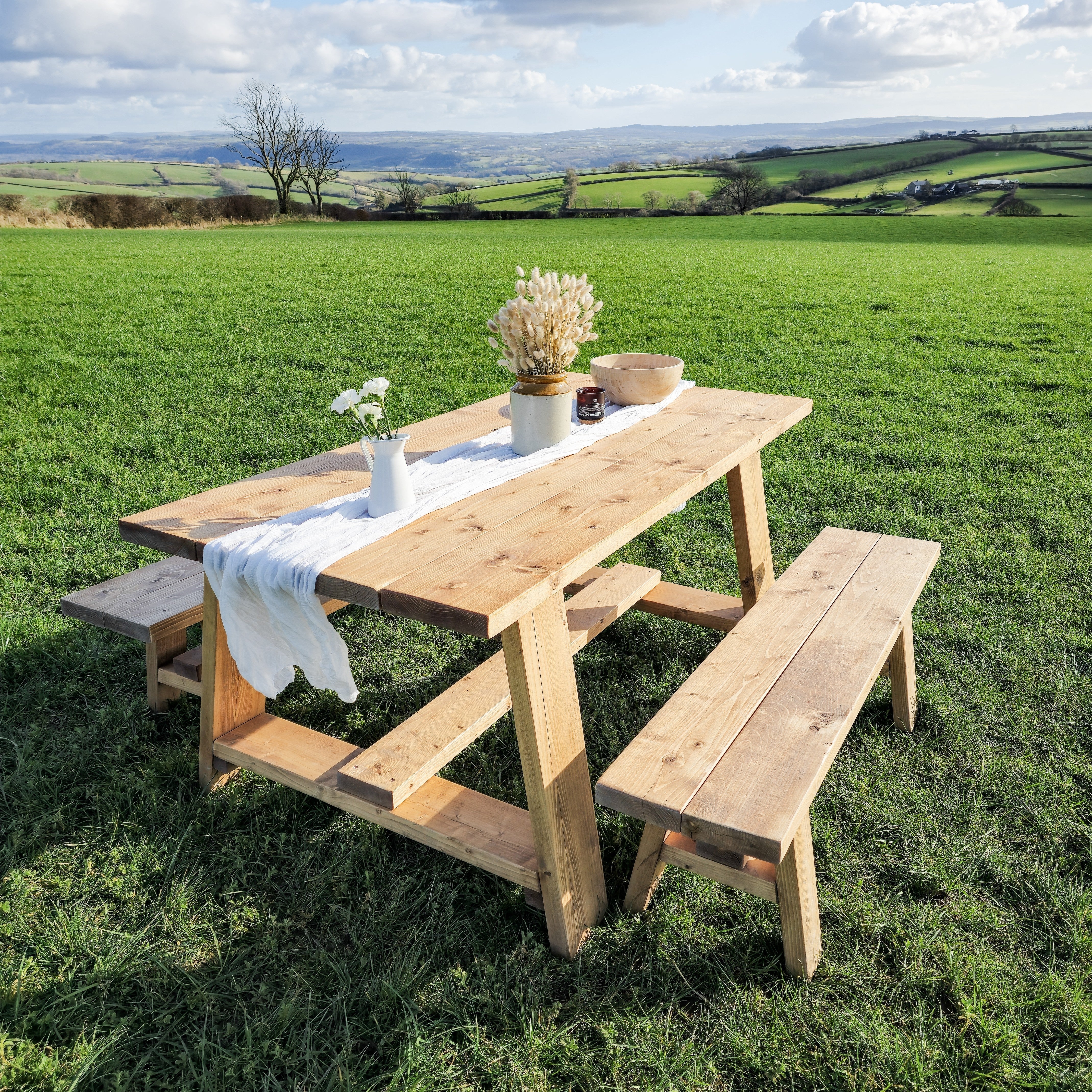 Light-colored wooden farmhouse outdoor table with timber legs and attached benches on grassy field showing table length