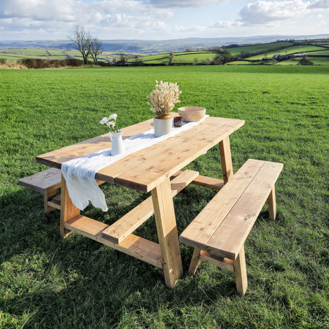 Light-colored wooden farmhouse outdoor table with timber legs and attached benches on grassy field showing table length