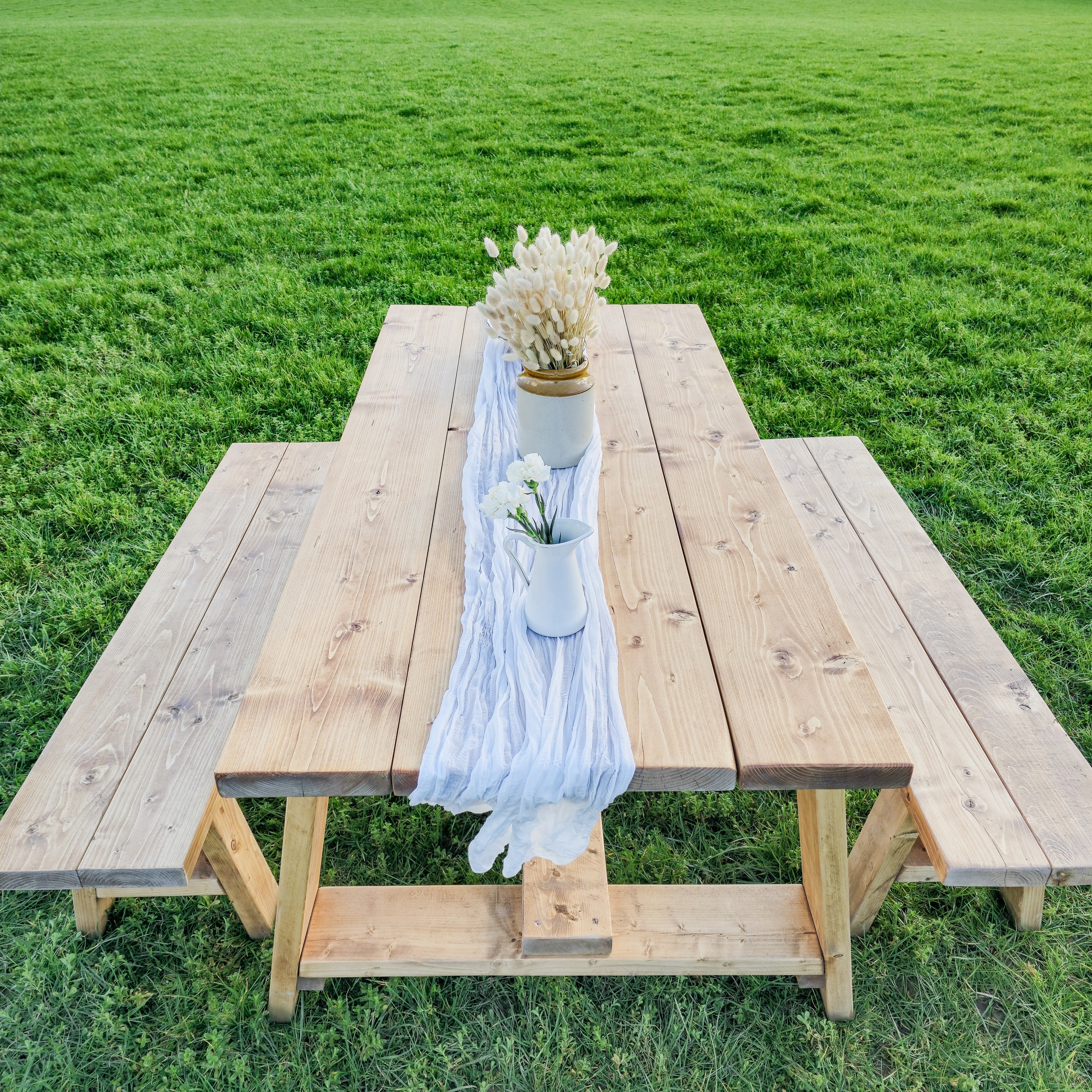 Light-colored wooden farmhouse outdoor table with timber legs and bench featuring a table length runner with vases