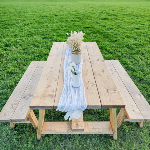 Light-colored wooden farmhouse outdoor table with timber legs and bench featuring a table length runner with vases