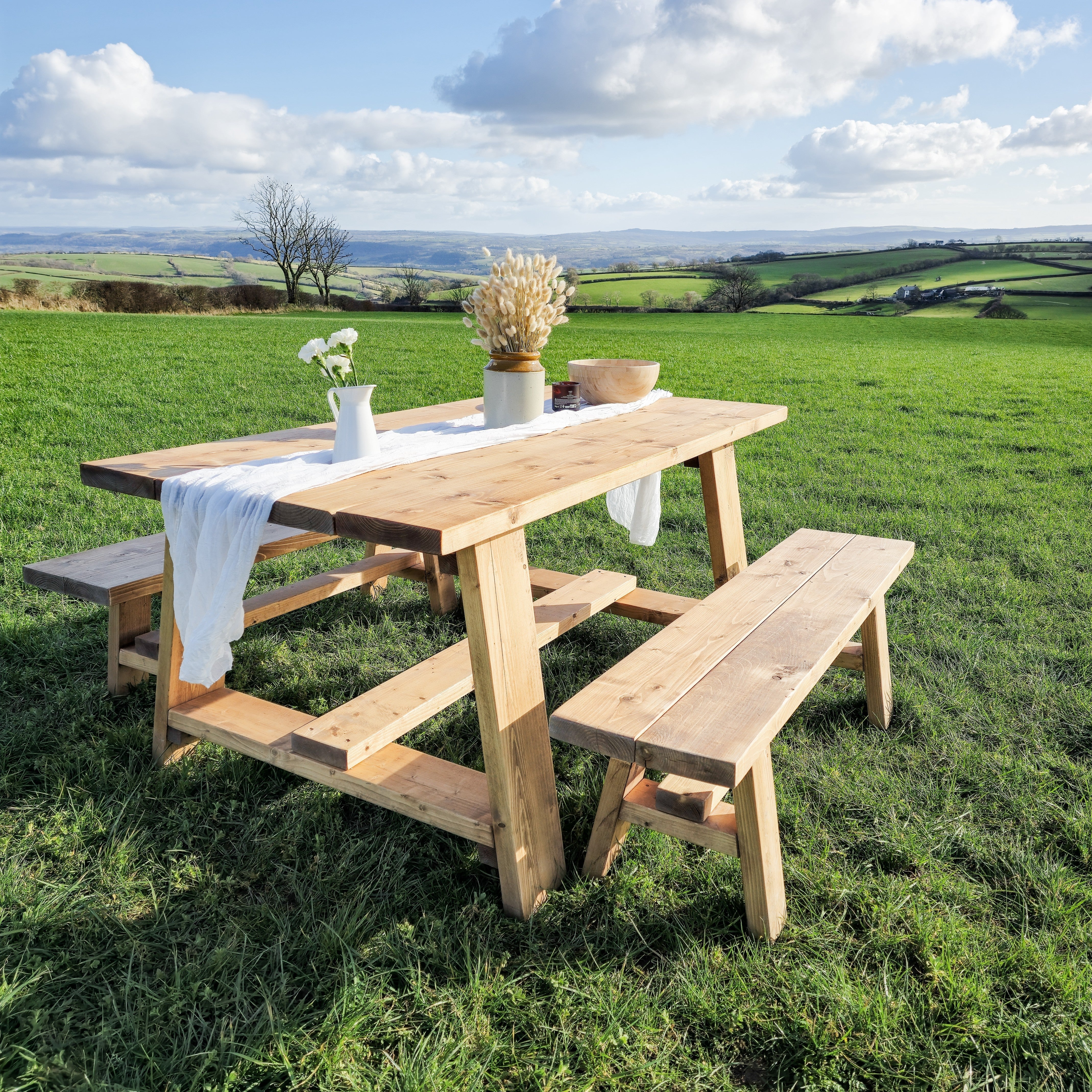 Light-colored wooden farmhouse outdoor table with timber legs and attached bench set on grassy field showing table length