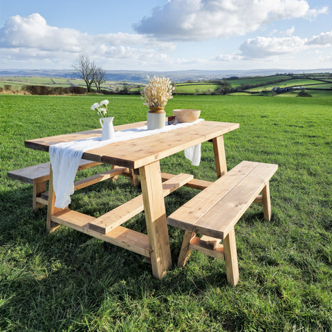 Light-colored wooden farmhouse outdoor table with timber legs and attached bench set on grassy field showing table length
