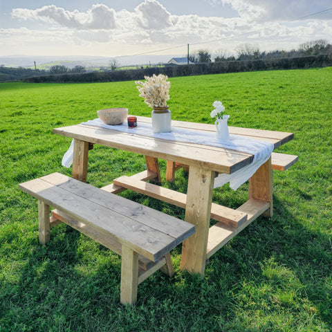 Light-colored wooden farmhouse outdoor table with timber legs and attached benches on grassy field showing table length