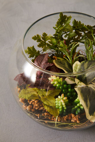 A clear glass spherical fiori large terranium with green and variegated artificial plants moss and pebbles inside