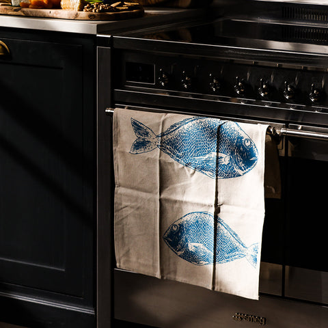 Beige linen tea towel with blue fish repeat design hanging from stove rack for kitchen use