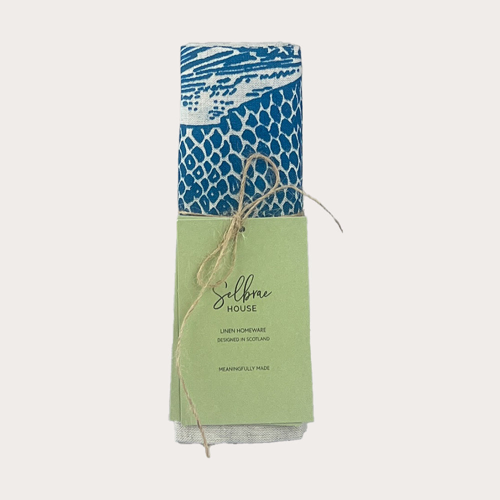 A rolled blue and white fish repeat linen tea towel tied with twine in a light green branded sleeve