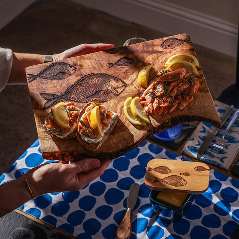 Hand-carved olive wood chopping board with rustic edge and engraved fish designs in warm brown tones
