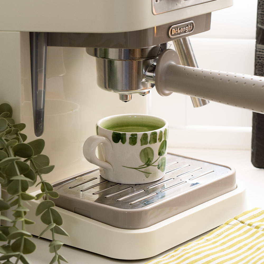 White ceramic espresso cup with hand painted green leafy pattern under a coffee machine for easy use