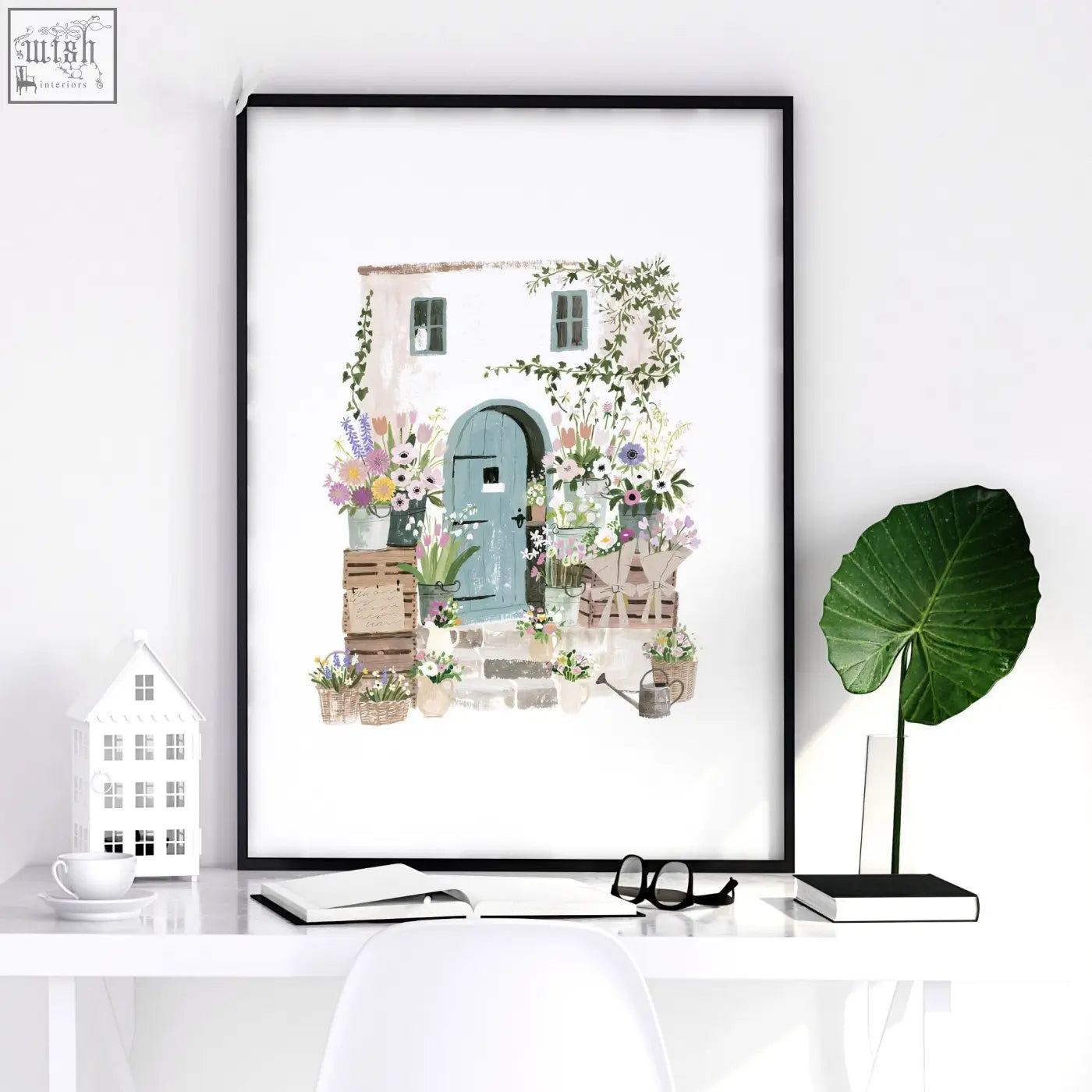 A framed watercolor illustrated print of a charming country cottage with a teal door and floral art in a black frame
