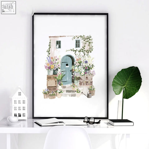 A framed watercolor illustrated print of a charming country cottage with a teal door and floral art in a black frame