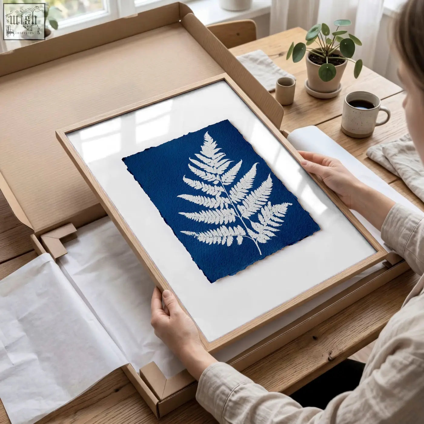 A framed floral art wall cyanotype print with a white fern leaf on deep blue paper in a light wood frame