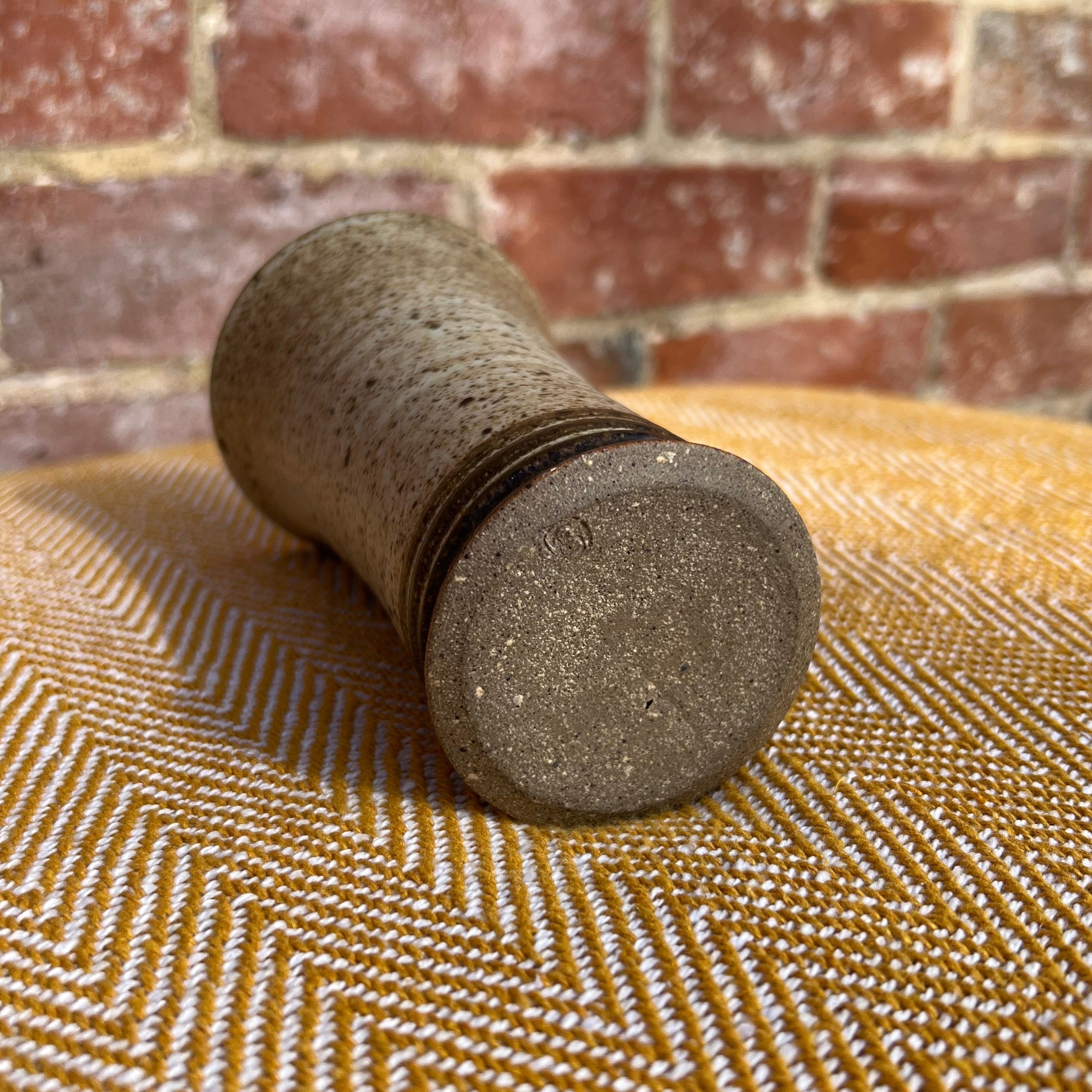 A rustic speckled gray ceramic pepper mill with textured glaze shown with a floral ceramic vase in the background