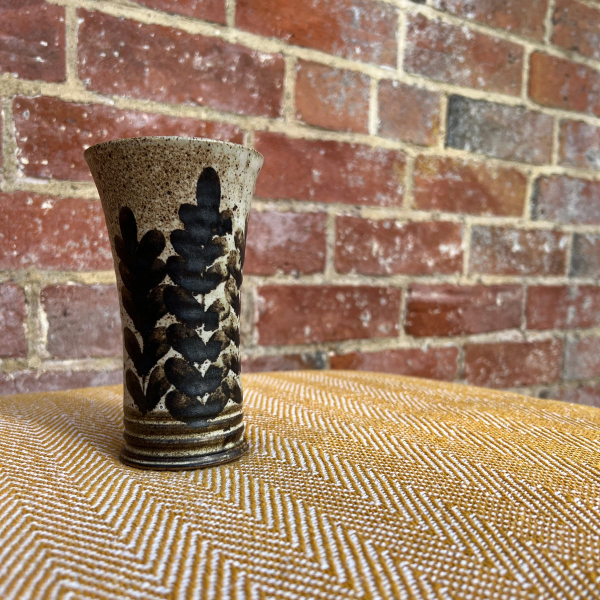 A floral ceramic vase with speckled beige glaze and black leafy patterns on a textured yellow surface
