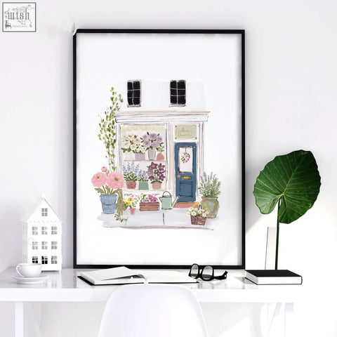 A framed watercolor flower art wall print of a pastel pink blue and white flower shop in a black frame