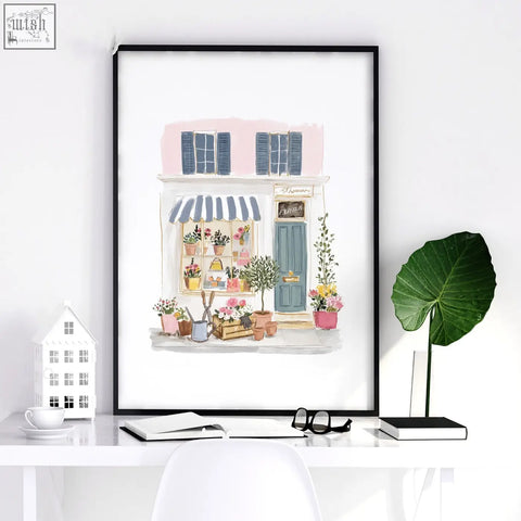 A framed watercolor flower art print of a charming flower shop with blue shutters and pastel pink accents