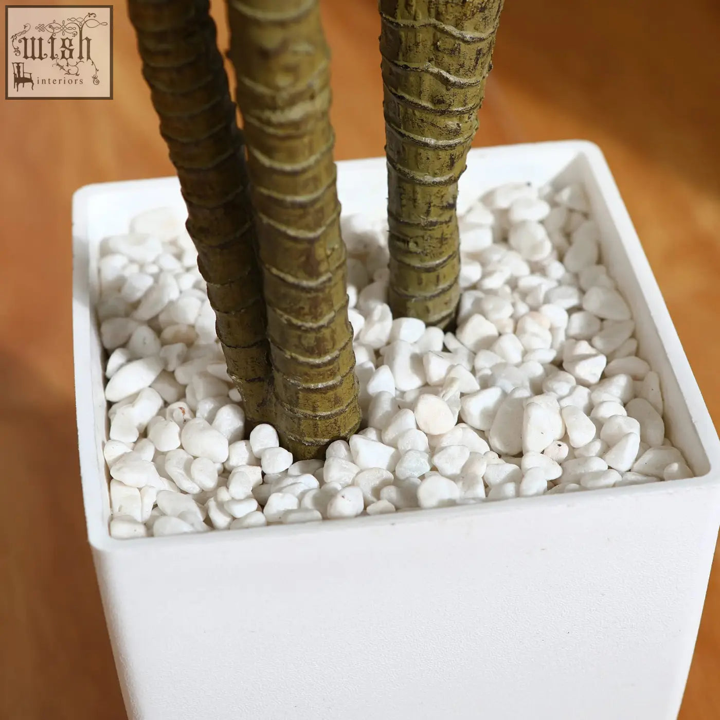 White square planter with white pebbles holding three artificial dracaena tree trunks for indoor decor