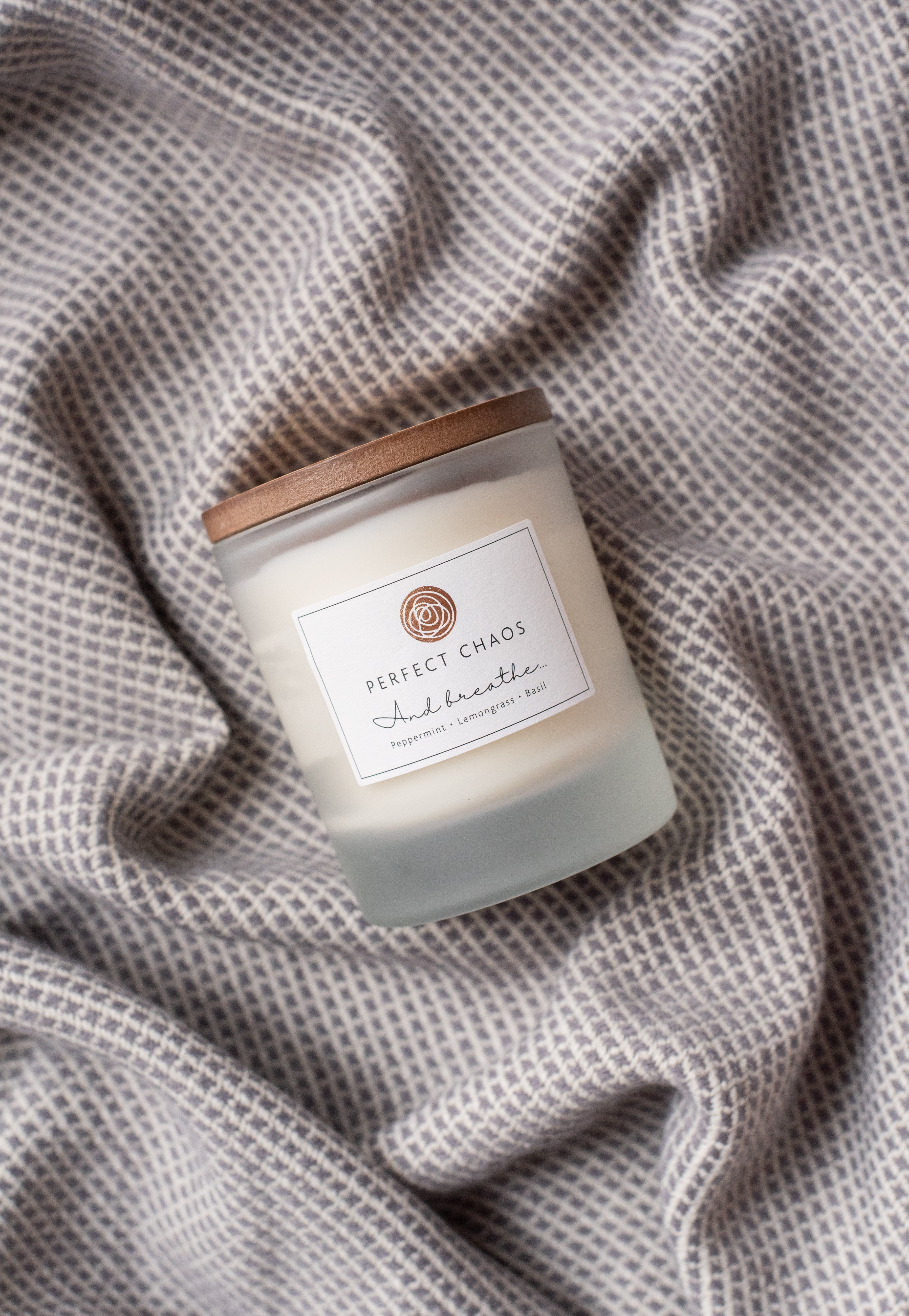A frosted glass candle jar with wooden lid labeled perfect chaos and breathe scented with peppermint lemongrass basil essential oils and hours burn time
